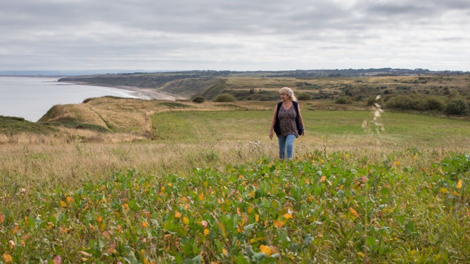 A women walking in a field next to the coast, with fields behind and the sea and sea cliffs of to one side